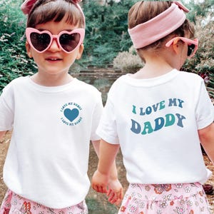 May include: Two children wearing white t-shirts with the text "I LOVE MY DADDY". One shirt has a heart graphic. The other shirt has the text on the back. Both children wear pink heart-shaped sunglasses and headbands.