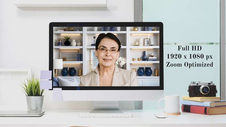 May include: A woman on a video call on a computer monitor. The monitor is on a white desk with a white mug, a camera, and books. The text on the monitor reads "Full HD 1920 x 1080 px Zoom Optimized."