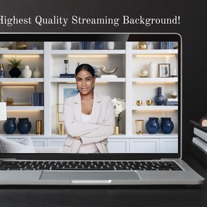 May include: A laptop computer displaying a woman with her arms crossed in front of a white bookshelf with blue and gold decorative items. The laptop is on a black surface with a black coffee mug and a stack of books.