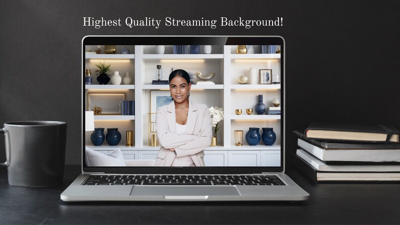 May include: A laptop computer displaying a woman with her arms crossed in front of a white bookshelf with blue and gold decorative items. The laptop is on a black surface with a black coffee mug and a stack of books.