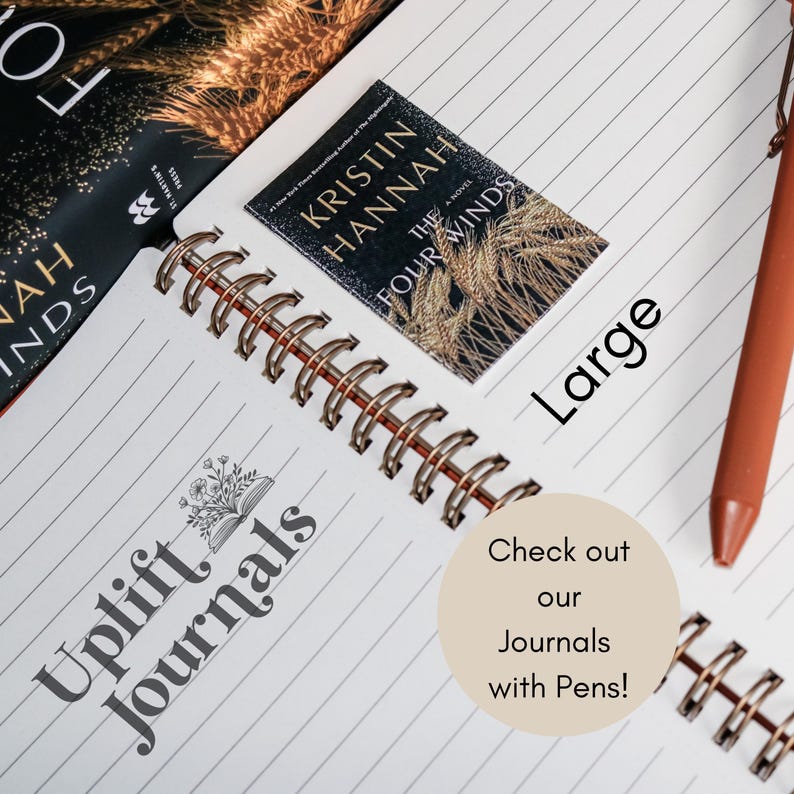 May include: A close-up of a spiral bound journal with the words "Uplift Journals" printed on the cover. The journal is open to a blank page with a book-shaped design and a quote from the book "The Four Winds" by Kristin Hannah. The text "Large" is written on the page. A brown pen is lying on the page. A sticker with the text "Check out our Journals with Pens!" is on the page.