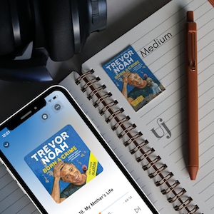 May include: A close-up of a smartphone screen displaying the audiobook "Trevor Noah: Born a Crime" on the Audible app. The screen shows the title of the audiobook, the author's name, and the chapter "My Mother's Life".