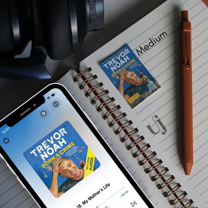 May include: A close-up of a smartphone screen displaying the audiobook "Trevor Noah: Born a Crime" on the Audible app. The screen shows the title of the audiobook, the author's name, and the chapter "My Mother's Life".