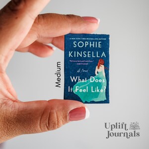 May include: A paperback book cover with a blue background and a woman in a turquoise dress. The title of the book is "What Does It Feel Like?" by Sophie Kinsella.