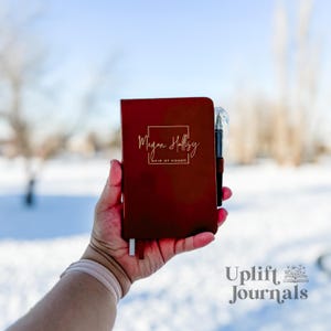 以下が含まれることがあります： 「Megan Halsey Maid of Honor」と金色の文字で刻まれた、えんじ色の革製のジャーナル。ジャーナルは手に持たれており、横にペンが置かれている。背景はぼやけており、雪景色が映っている。