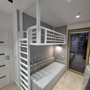 May include: A white metal loft bed frame with a built-in vertical ladder and safety railings, positioned directly above a light gray upholstered sofa. The structure features a clean, rectangular design with horizontal bars for the bed enclosure.