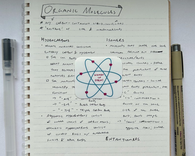 May include: A notebook page with handwritten notes about organic molecules, including a definition of hydrocarbons and a section on isomers. A sticker with the text "Women in STEM!" is placed on the page.