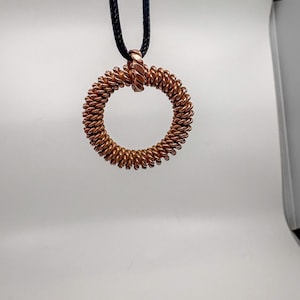 May include: A circular pendant crafted from coiled copper wire, featuring a textured, twisted metal surface. The pendant is suspended from a simple black cord, creating a contrast between the metallic copper tones and the dark cord material.