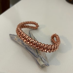 Può includere: Un bracciale a polsino color rame con un design a spirale. Il bracciale è composto da due fili paralleli di filo avvolto. Il bracciale è appoggiato su una pietra blu e bianca.