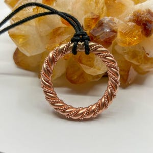 May include: A copper-colored pendant necklace featuring a circular, braided design. The pendant hangs from a black cord. The background is a cluster of golden-orange crystals, providing a contrasting backdrop to the jewelry.