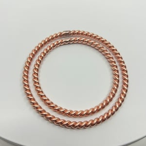 May include: Two copper-colored, twisted metal bangles. The bangles are circular and lie on a white surface. The twisted design gives the bangles a rope-like appearance. The metal has a warm, reddish-brown hue.