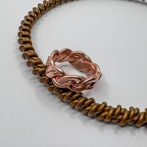 May include: A copper-colored braided ring rests on a coiled copper-colored wire. The ring has a woven design, and the wire is arranged in a circular shape. The background is white.