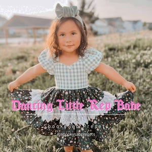 May include: A young girl wearing a green and white gingham dress with a floral print skirt. The dress has a matching headband and the text "Dancing Little Rep Babe" is across the bottom of the image. The dress is perfect for a special occasion.
