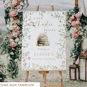 May include: A rectangular welcome sign on a wooden easel featuring a floral border with green leaves and pink, white, and yellow flowers. The text reads A NEW CHAPTER BEGINS, an illustration of stacked books, and WELCOME TO ELOISE'S BRIDAL SHOWER.