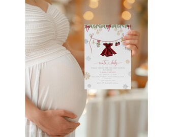 May include: A Christmas-themed baby shower invitation card held by a person. The card features a red dress and baby shoes hanging on a clothesline, with the words "santa & baby" in cursive. Gold and white snowflakes and greenery decorate the top.
