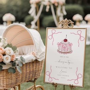 Puede incluir: Una escena de baby shower con un cochecito de mimbre vintage y arreglos florales, junto a un letrero. El letrero, en un marco dorado, dice "She is the cherry on top" con una ilustración de pastel y "Welcome to Emma's Baby Shower".