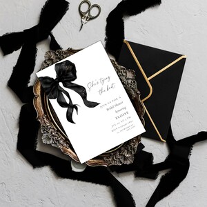 Peut inclure: Une invitation à une fête prénuptiale avec le texte "She's tying the knot" et des détails. L'invitation est sur une carte blanche avec un motif de nœud noir, posée sur un plateau décoratif doré, avec une enveloppe et un ruban noirs.