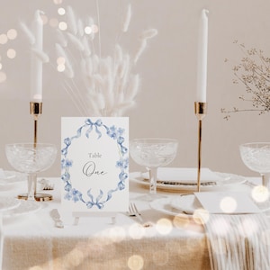 Könnte beinhalten: Eine weiße Karte mit einem blauen Aquarellkranz und dem Text "Tisch Eins" auf einem Tischgedeck mit Kerzen und Gläsern.