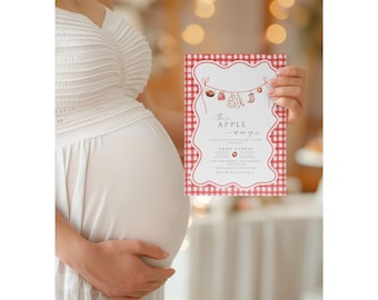 May include: A baby shower invitation with a red and white gingham border. The invitation features the text "This Apple of our eye" with decorative elements of apples and baby clothes. The invitation is held by a person wearing a white dress.