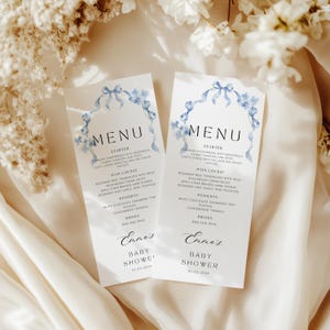 May include: Two light blue and white menus with a watercolor design of blue ribbons and flowers. The menus are for a baby shower and list the starter, main course, desserts, and drinks. The text on the menus reads "Emma's Baby Shower 01-03-2024".
