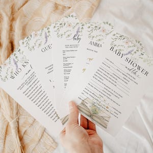 May include: A collection of baby shower game cards with floral and lavender accents. The cards feature various game titles, including "Baby Shower," "Who Knows Mom?," and "Guess the Baby." The design is soft and inviting, perfect for a baby shower.