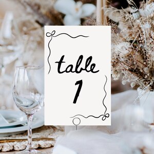 Puede incluir: Tarjeta de número de mesa blanca con caligrafía negra que dice "mesa 1" con un borde decorativo.