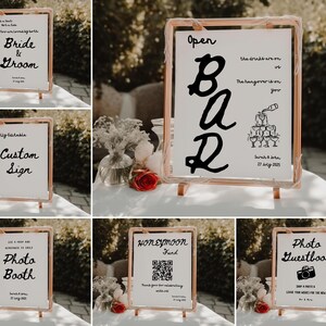 May include: Six wedding signs with black calligraphy on white backgrounds. The signs are on wooden easels and include the text "Bride & Groom", "Custom Sign", "Open Bar", "Photo Booth", "Honeymoon Fund", and "Photo Guestbook".