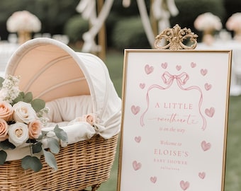 May include: A baby shower scene featuring a vintage wicker baby carriage with a peach-colored interior and floral arrangement. A framed sign with pink hearts and text that reads "A Little Sweetheart is on the way" is displayed.
