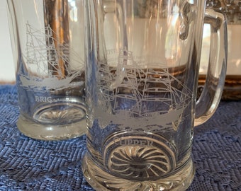 May include: Two clear glass beer mugs with etched images of sailing ships. The mug on the right has the word "CLIPPER" etched on the bottom. The mug on the left has the word "BRIG" etched on the bottom.