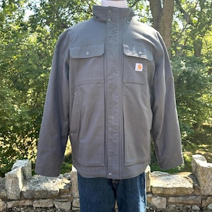 May include: A gray work jacket with multiple pockets and a high collar, displayed on a mannequin. The jacket features a small orange and white logo on the front and is paired with blue jeans.