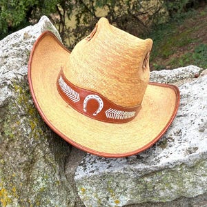 May include: A straw cowboy hat with a brown leather band featuring a white horseshoe and arrow design. The hat has a wide brim and a tall crown. The hat is resting on a stone wall.