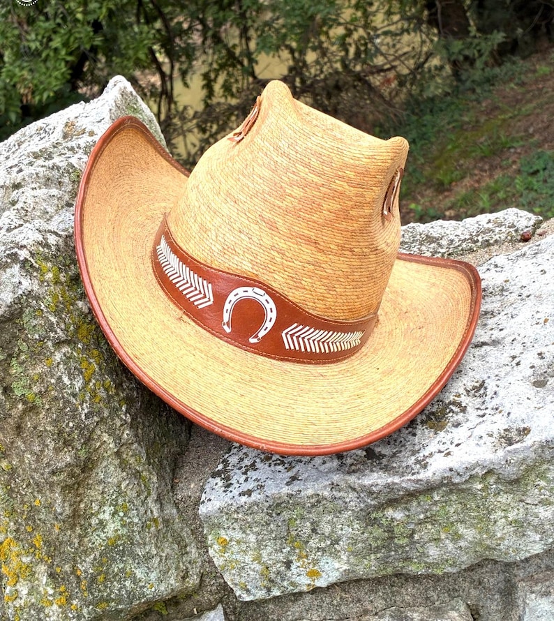 May include: A straw cowboy hat with a brown leather band featuring a white horseshoe and arrow design. The hat has a wide brim and a tall crown. The hat is resting on a stone wall.