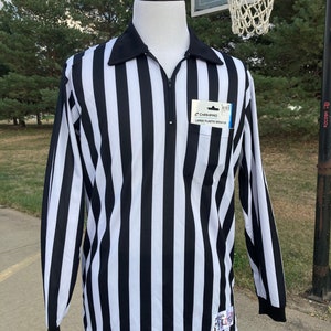 May include: Black and white striped referee shirt with a zipper and a pocket on the left side. The shirt has a Champro label with the text "Large Plastic Whistle".