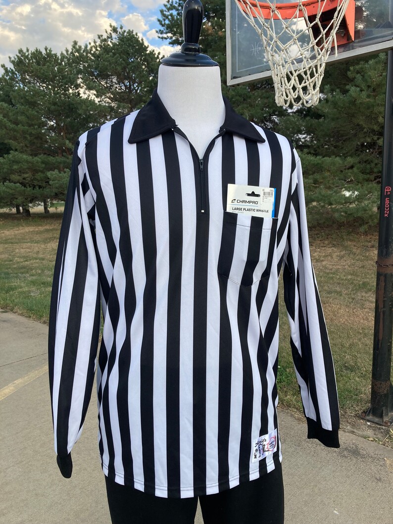 May include: Black and white striped referee shirt with a zipper and a pocket on the left side. The shirt has a Champro label with the text "Large Plastic Whistle".