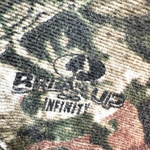 May include: Close-up of a camouflage pattern fabric with the text "Break Up Infinity" in black. The fabric features a blend of green, brown, and tan colors, with a textured appearance. The Mossy Oak logo is also visible.