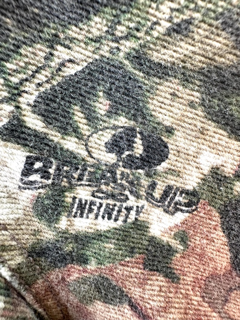 May include: Close-up of a camouflage pattern fabric with the text "Break Up Infinity" in black. The fabric features a blend of green, brown, and tan colors, with a textured appearance. The Mossy Oak logo is also visible.