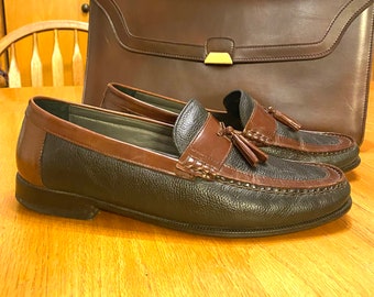 May include: A pair of brown and black leather tassel loafers. The loafers have a brown leather upper with a black leather toe cap and a tassel on the top.