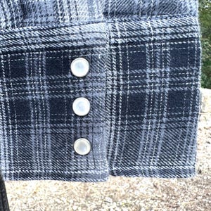 May include: Close-up of a shirt cuff with a blue and gray plaid pattern. The cuff features three silver-colored snap closures. The fabric has a textured appearance.