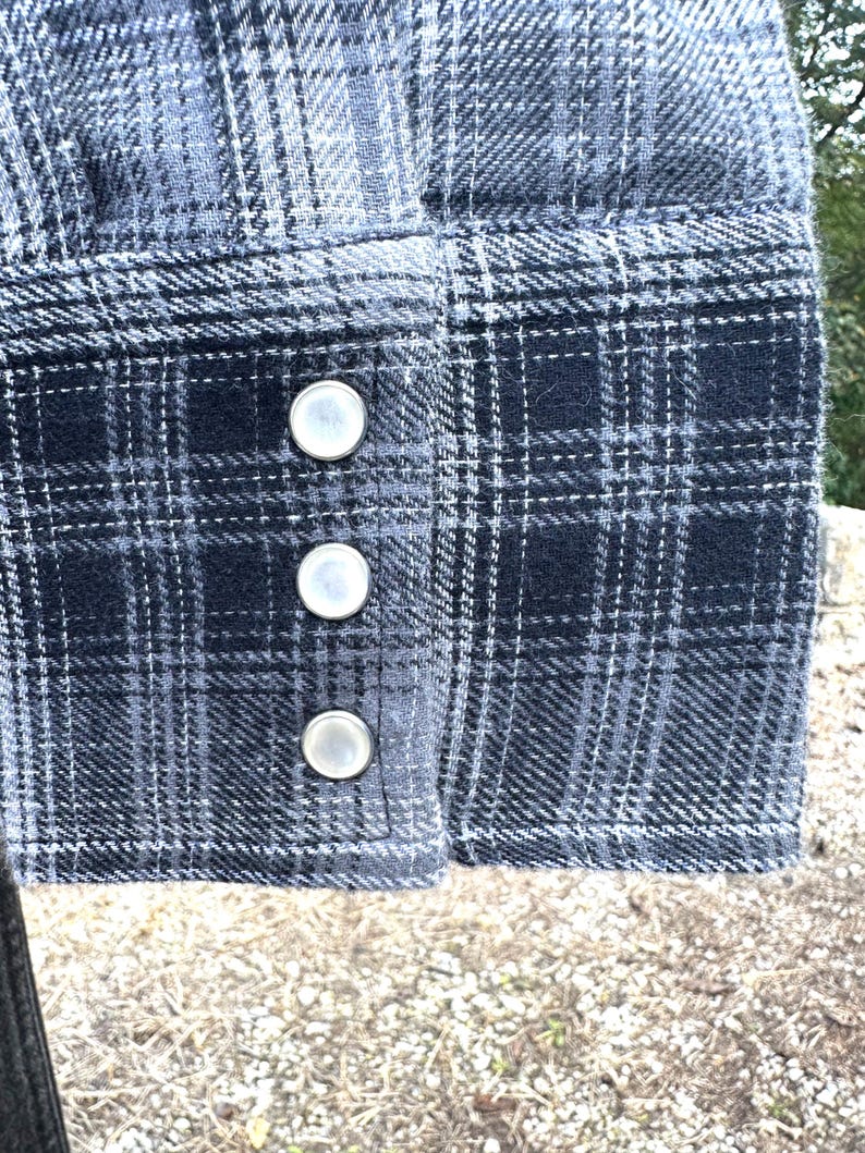 May include: Close-up of a shirt cuff with a blue and gray plaid pattern. The cuff features three silver-colored snap closures. The fabric has a textured appearance.