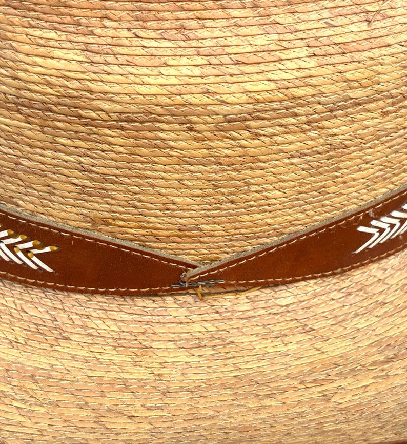 May include: Close-up of a straw hat with a brown leather band. The band is decorated with white and yellow arrow-like designs. The hat has a woven texture and a natural colour.