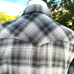 May include: A gray and white plaid shirt, viewed from the back, is displayed on a mannequin. The shirt features a classic western yoke design and a button-down collar. The plaid pattern is a mix of light and dark gray tones.