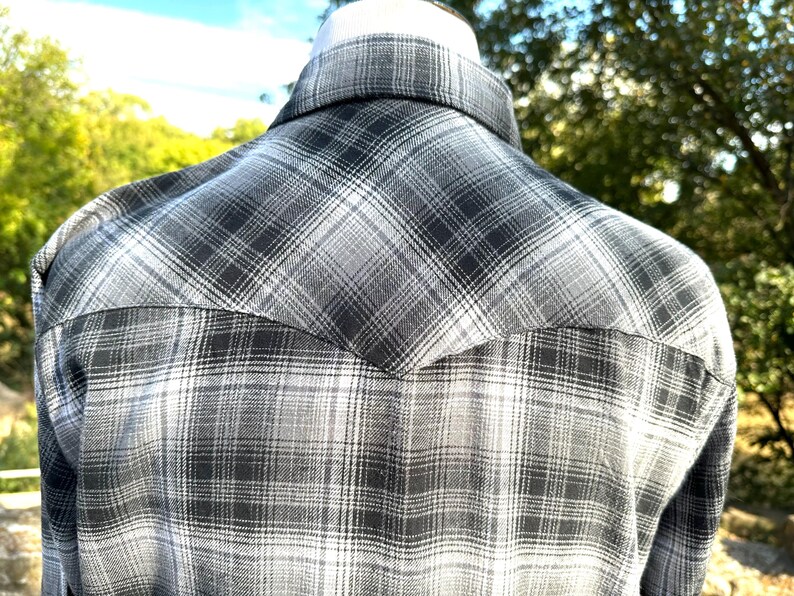 May include: A gray and white plaid shirt, viewed from the back, is displayed on a mannequin. The shirt features a classic western yoke design and a button-down collar. The plaid pattern is a mix of light and dark gray tones.