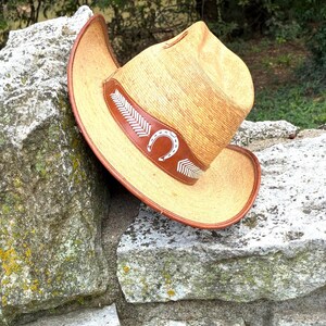 May include: A straw cowboy hat with a brown leather band featuring a white horseshoe and arrow design. The hat has a wide brim and is resting on a stone wall. The hat is a light tan colour.