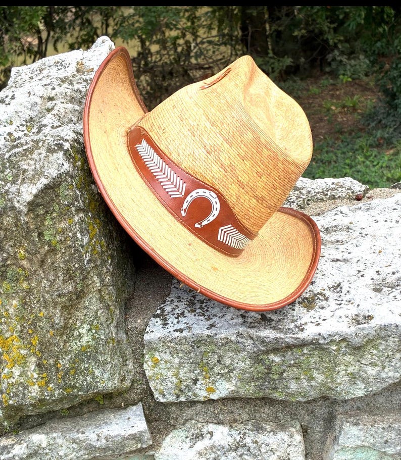 May include: A straw cowboy hat with a brown leather band featuring a white horseshoe and arrow design. The hat has a wide brim and is resting on a stone wall. The hat is a light tan colour.