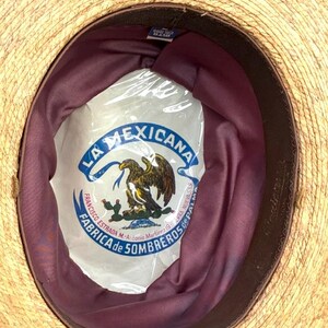 May include: A straw hat with a dark brown brim and a burgundy interior. The hat's interior features a white label with the text "LA MEXICANA" and an eagle emblem. The hat is likely a traditional sombrero.