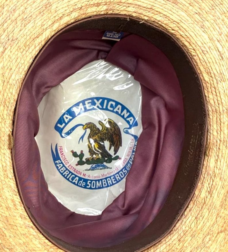 May include: A straw hat with a dark brown brim and a burgundy interior. The hat's interior features a white label with the text "LA MEXICANA" and an eagle emblem. The hat is likely a traditional sombrero.
