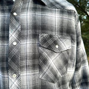 May include: A black, white, and gray plaid button-up shirt. The shirt has a classic western style with a pointed collar, two chest pockets, and pearl snap closures. The plaid pattern is a mix of dark and light shades.