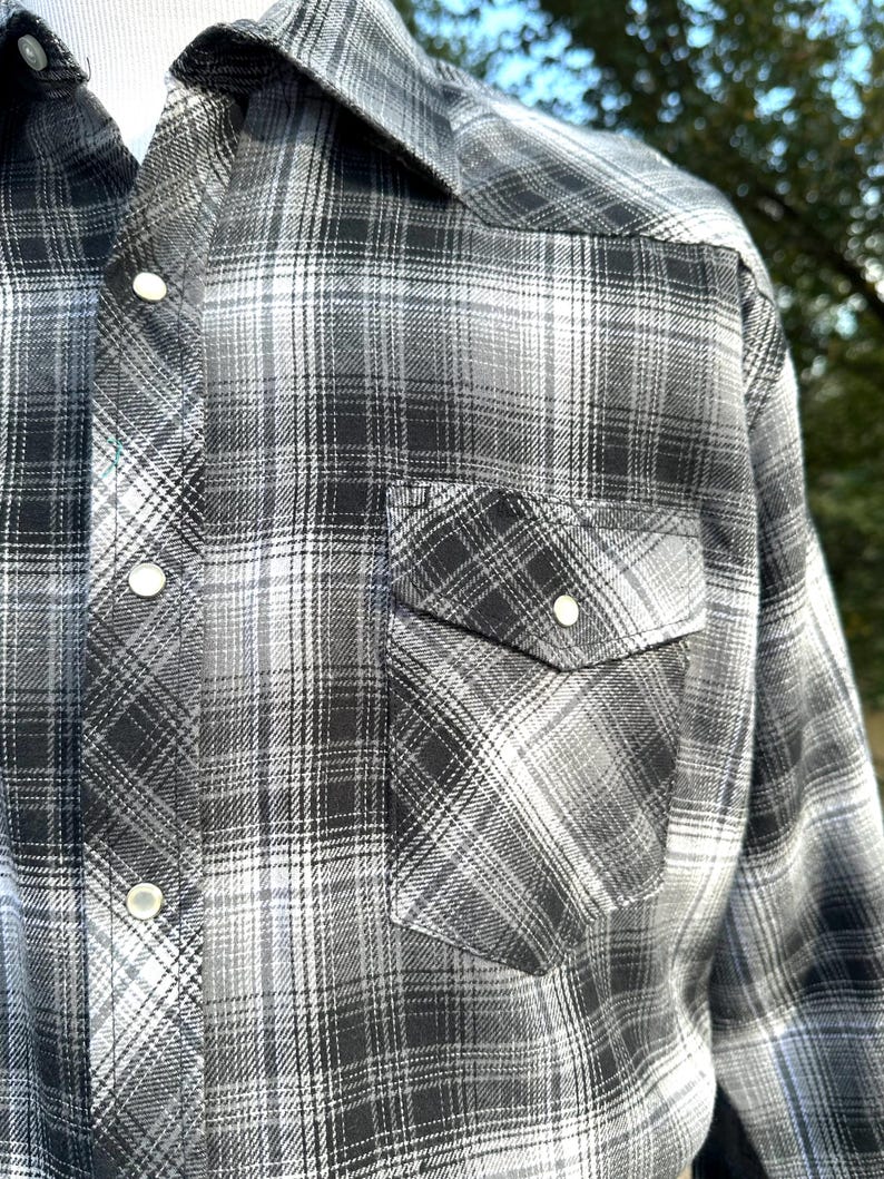 May include: A black, white, and gray plaid button-up shirt. The shirt has a classic western style with a pointed collar, two chest pockets, and pearl snap closures. The plaid pattern is a mix of dark and light shades.