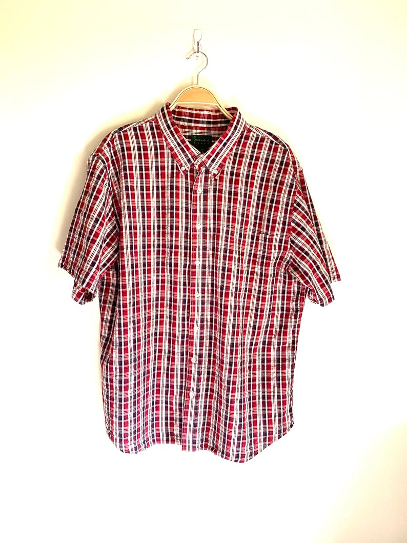May include: A red, white, and blue plaid short-sleeved button-down shirt. The shirt has a pocket on the left side.