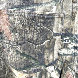 May include: Close-up of camouflage-patterned denim fabric, featuring shades of green, brown, and gray. The image highlights a back pocket and stitching details, showcasing the texture and design of the material. The overall theme is outdoor apparel.
